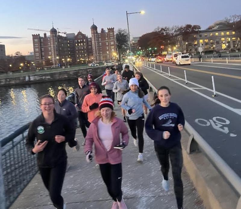A group of students running across the MIT bridge over the Charles River at dusk.