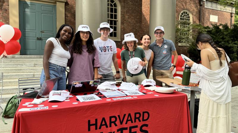 a group of students behind a table with a "HARVARD VOTES" banner. They are wearing white "VOTE" bucket hats and promoting voting. The table has a laptop, hats, badges, and informational materials. The scene is outdoors, likely on a university campus, and everyone is smiling, creating a positive atmosphere.