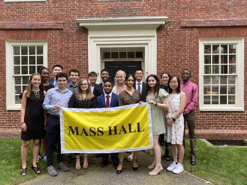 A group picture of residents of Massachusetts Hall holding a yellow banner that reads "Mass Hall" in front of a red brick building.