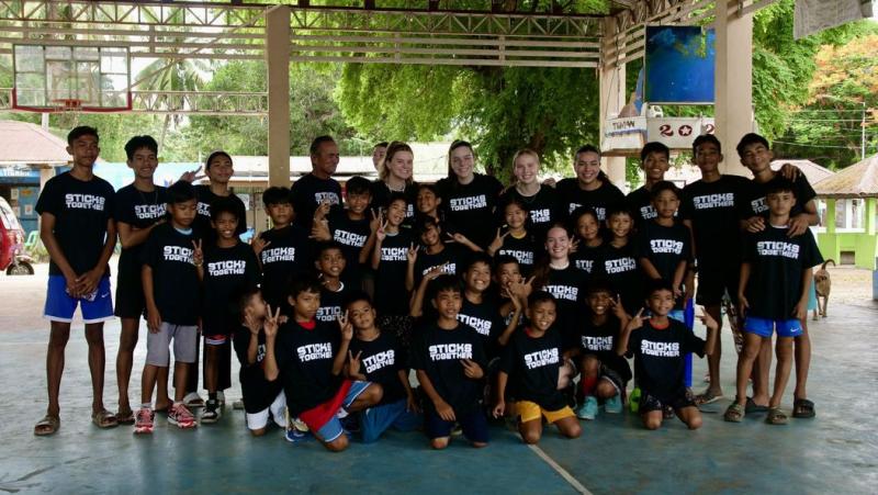 A large group of smiling children and adults wearing matching "Sticks Together" shirts on a basketball court.