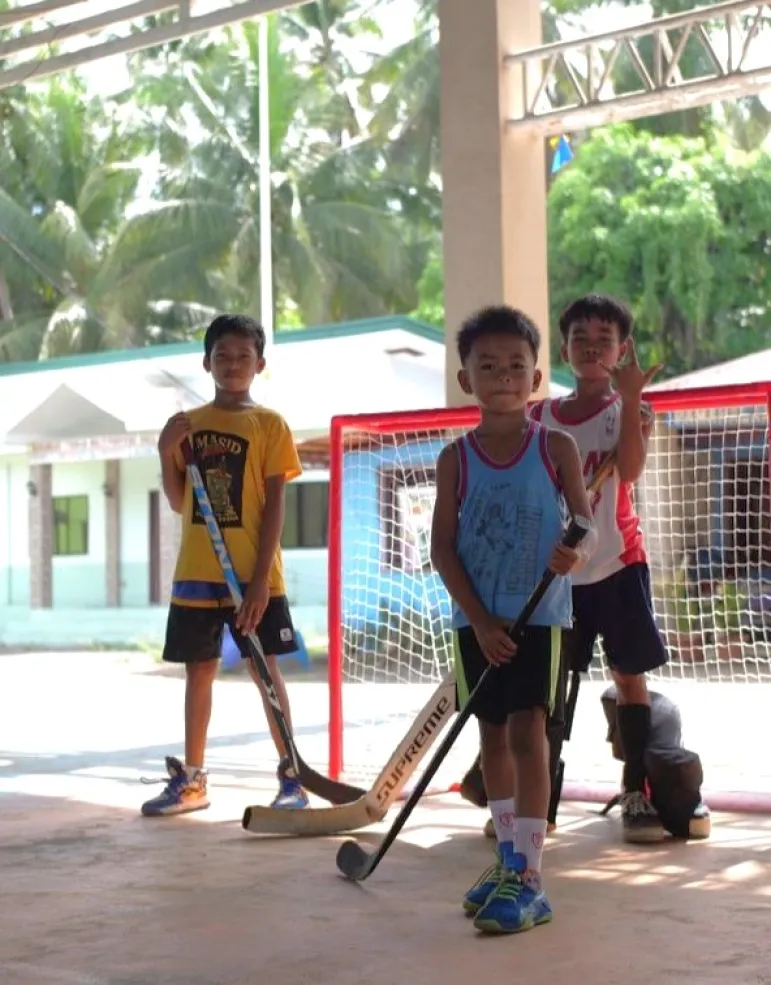 Three young children with hockey sticks stand in front of a net on an outdoor court
