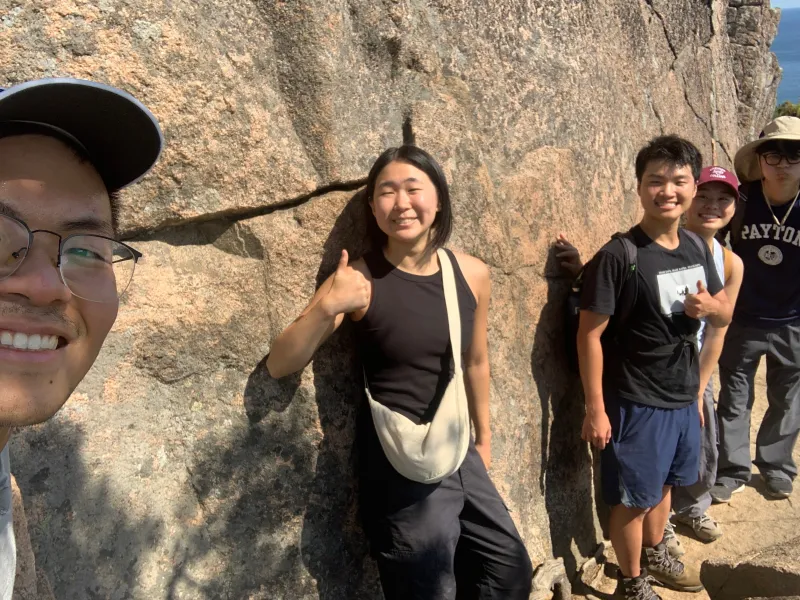 Selfie picture of Raymond and his friends clinging onto a stone wall while hiking the Beehive Trail at Acadia National Park.