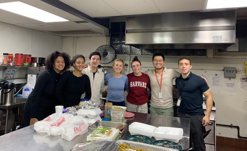 Picture of Raymond, his friends, and evening shelter volunteers smiling at camera in the kitchen of the Harvard Square Homeless Shelter.