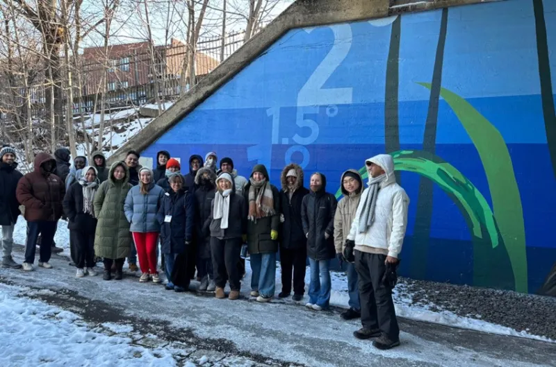 A group of people in winter clothing, standing on a snowy path beside a large, colorful mural with numbers and abstract designs. 