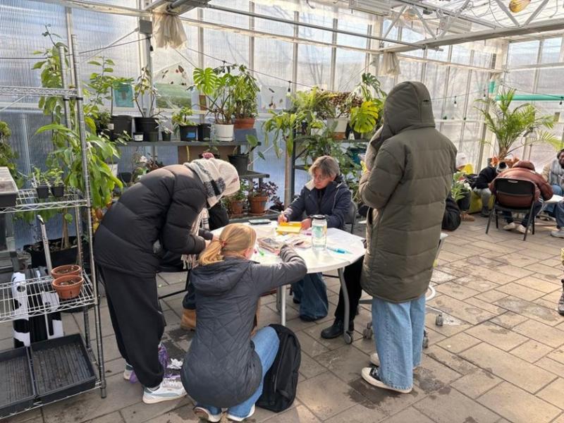 People gathered around a table inside a greenhouse filled with various plants. Some are seated while others are standing.