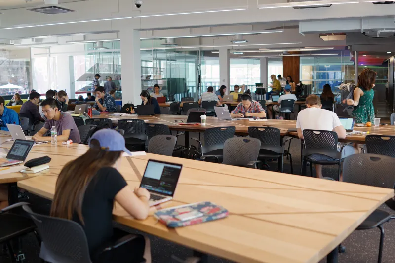 People studying and working on laptops in a large, bright workspace with long tables.