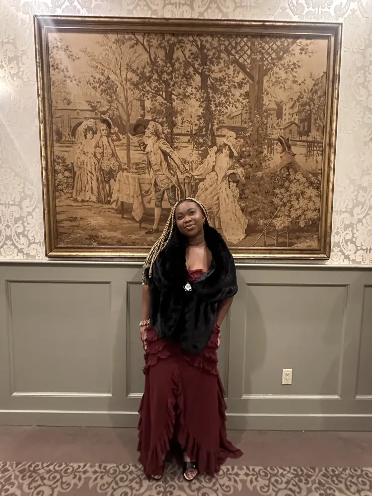 A picture of a black girl with blond box braids in a long red dress and a black fur coat over the dress. She is standing by the wall infront of a picture posing