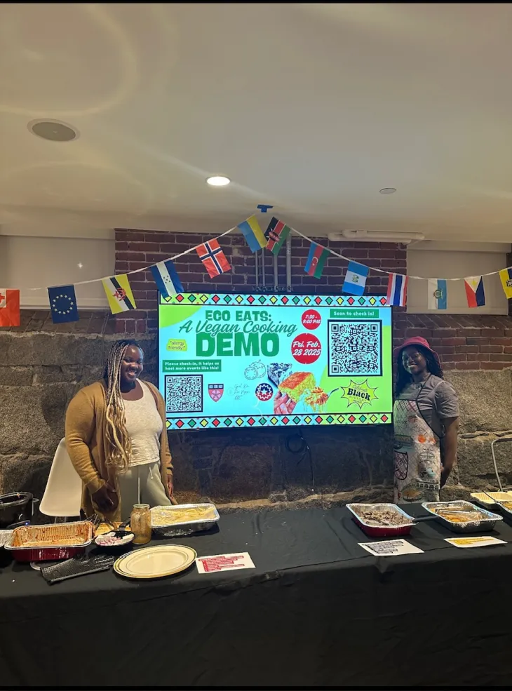Two black women standing infront of a screen labeled "Eco Eats: A Vegan Cooking Demo. There is also a table of food infront of them