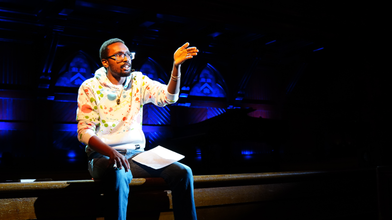 A man sitting on the edge of a stage speaking and pointing to the audience.