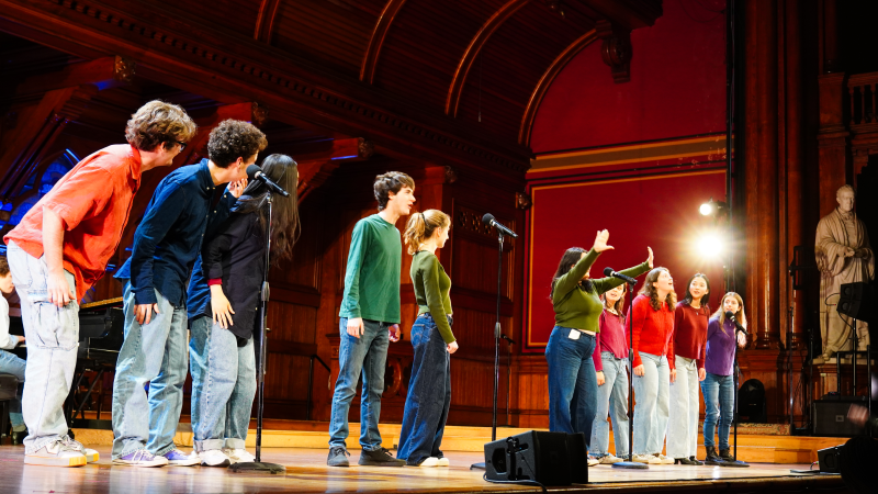 A group of performers on stage, singing into microphones, with a spotlight illuminating them.