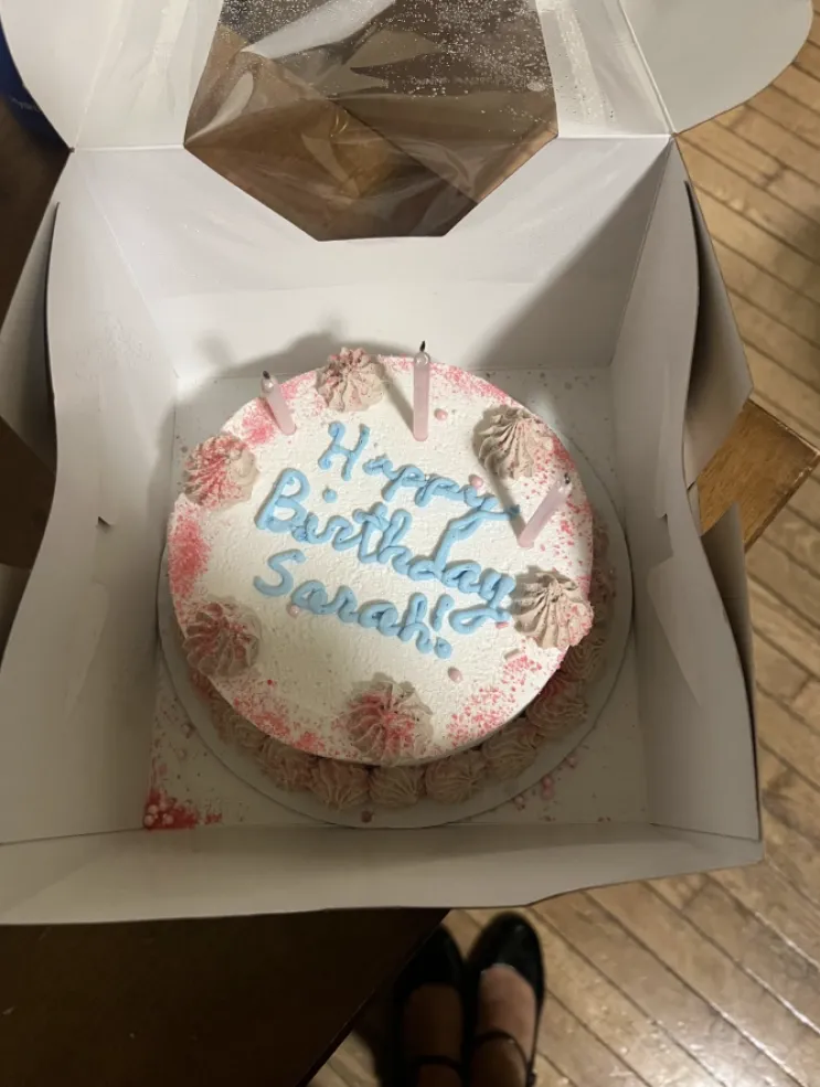 White birthday cake with pink sprinkles and blue writing with the words, "Happy Birthday Sarah!"