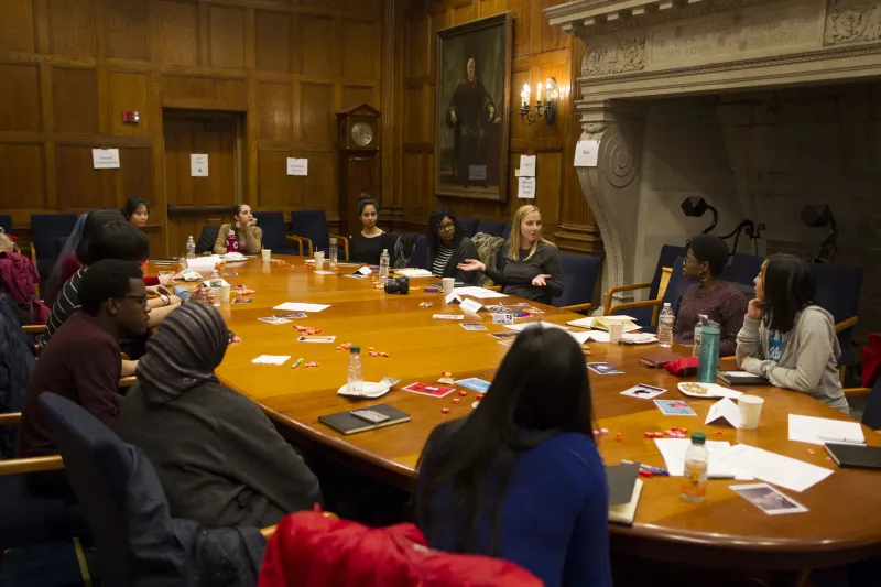 People having a discussion around a large wooden table in a wood-paneled room.