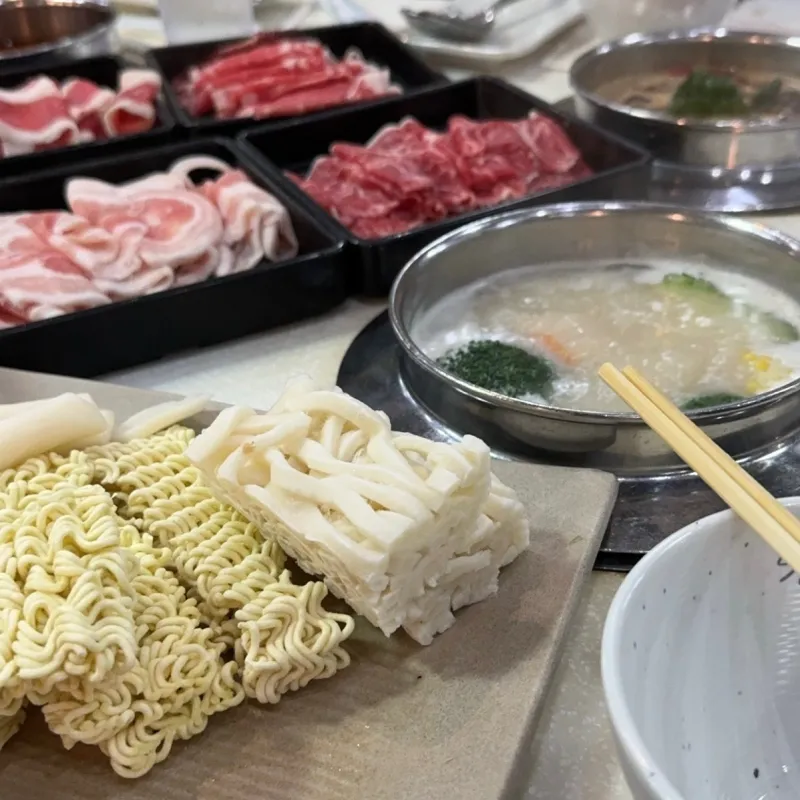  A picture of hot pot broth, a plate of noodles, and multiple plates of meat from Spring Shabu Shabu.