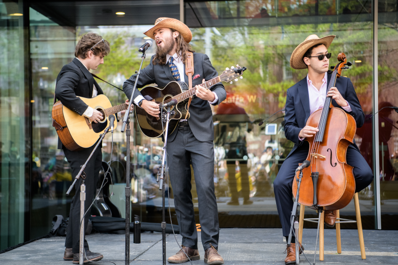 Three musicians in suits and cowboy hats perform outdoors, playing acoustic guitar, electric guitar, and cello.