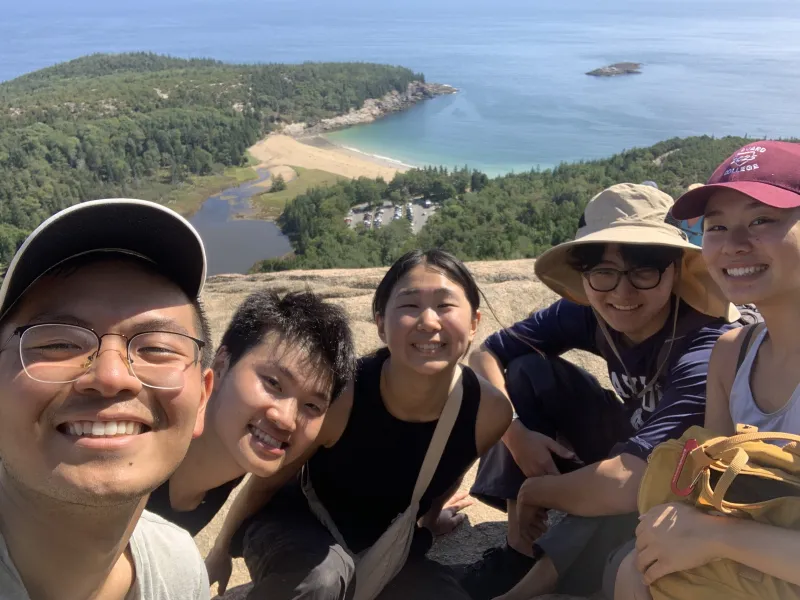A group picture of Raymond and his friends near the top of the Beehive Trail with Acadia National Park and the ocean horizon in the background.