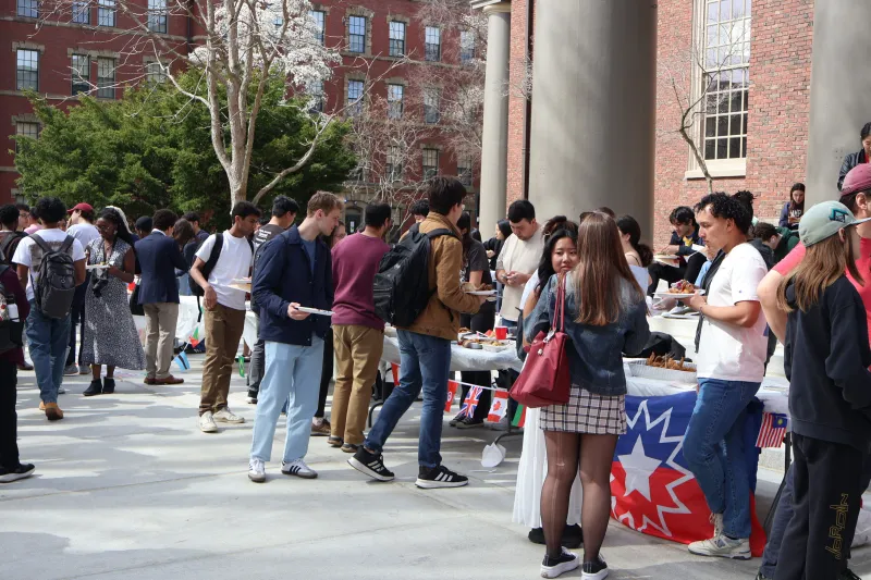 A picture of a bunch of students gathered on the steps of Memorial Church. They are walking around to different tables to try food. The tables also have flags from the represented countries hung on them