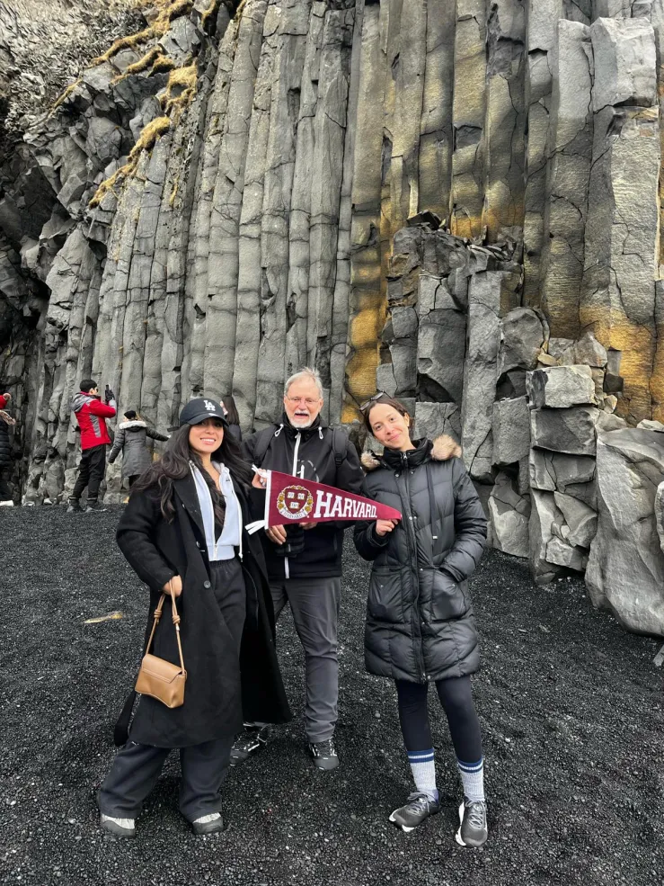 Three people posing for a photo in front of a rock wall, holding a Harvard flag