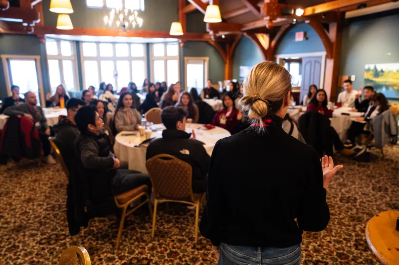 A woman speaks to a group of people seated at round tables in a warmly lit room with wooden beams and large windows.