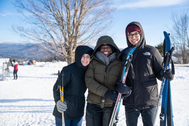 Three people in winter clothing smile while holding skis in the snowy outdoors.
