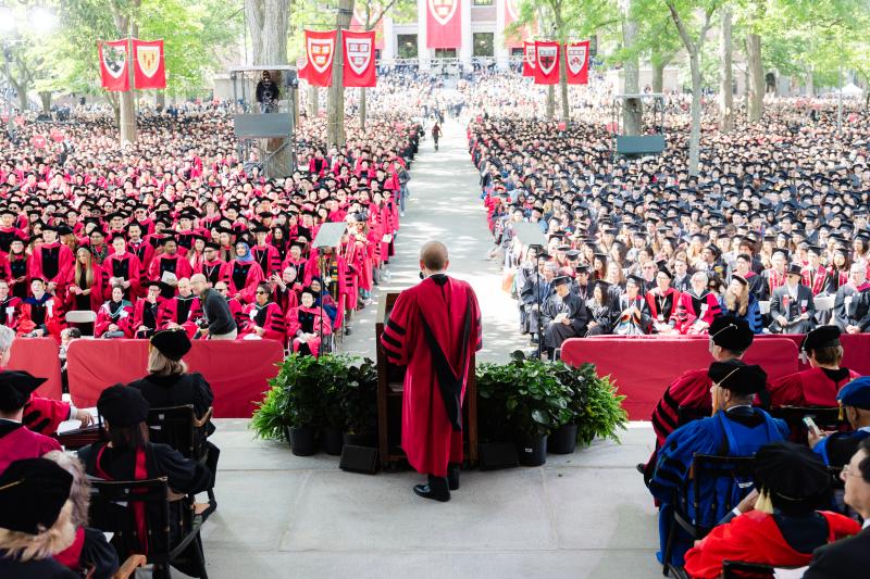 A speaker addresses graduates in caps and gowns at a large outdoor Harvard commencement ceremony.