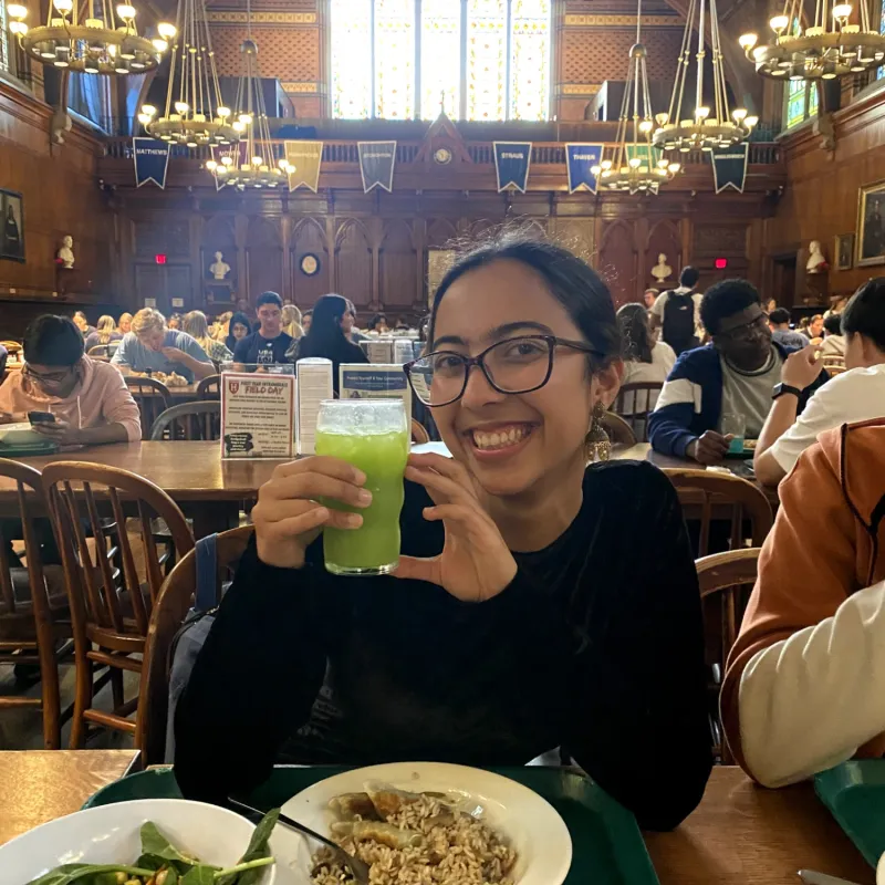 The author holding up a green drink she created, called Emerald Sparkle, at Annenberg, with grand chandeliers and freshman dorm flags in the background.