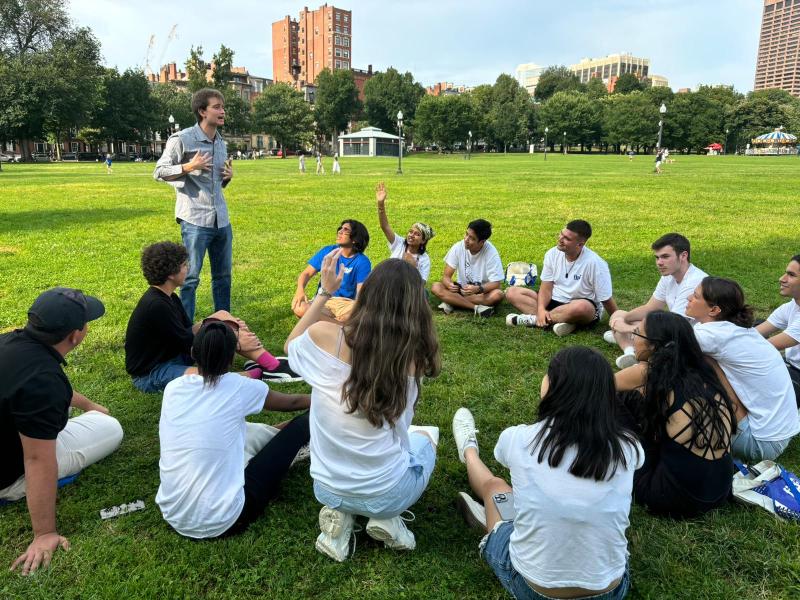 Several students sitting in a circle on grass while one person is standing
