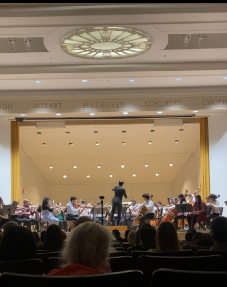 A group of Bach Society Orchestra members playing their instruments on stage, dressed in concert attire, captured mid-performance.
