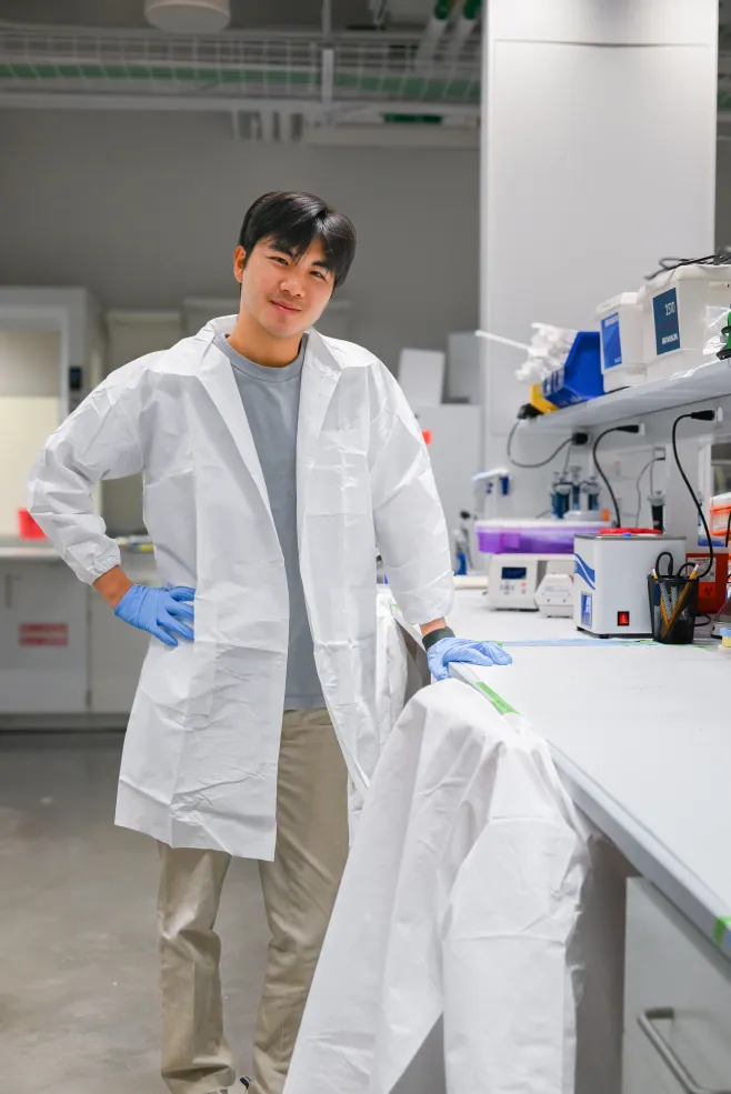 Sean stands against his lab station dressed in his white lab coat.
