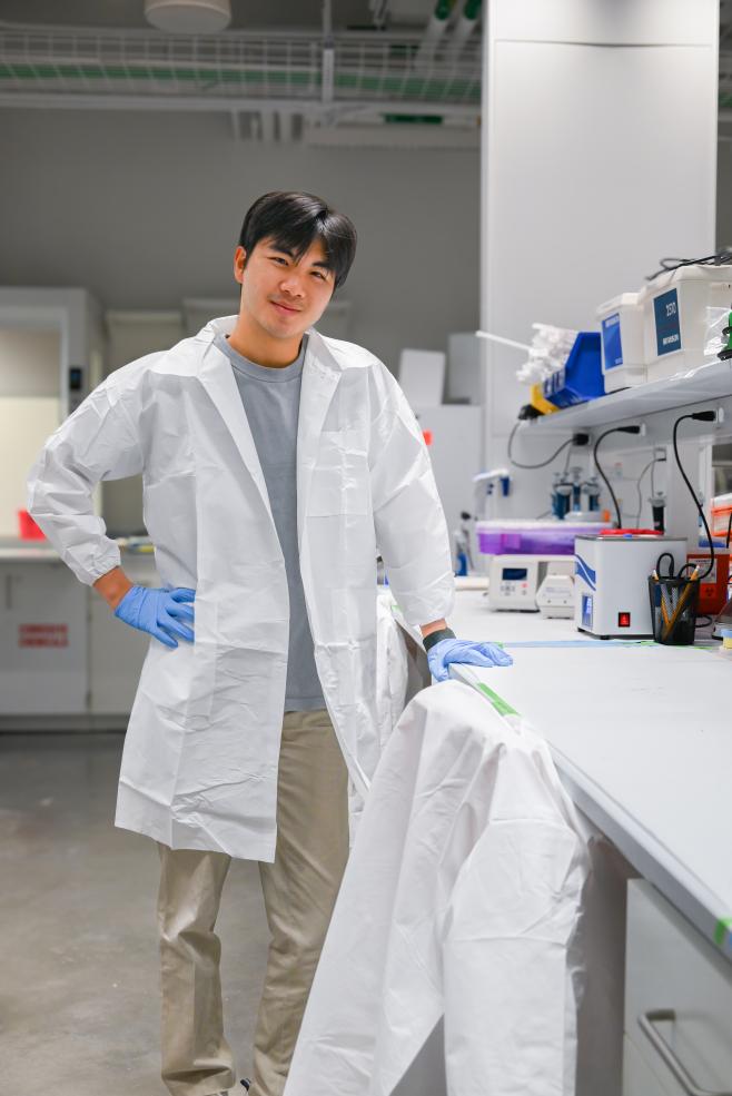 Sean stands against his lab station dressed in his white lab coat.
