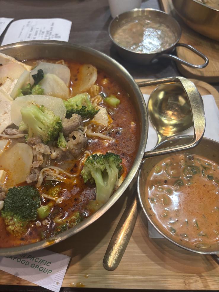 A medium sized metal pot is pictured. It contains spicy soup filled with broccoli, sliced beef, potatoes, etc.