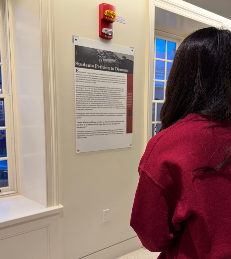 Person in a red sweater reading a wall display titled "Students Petition to Dename."