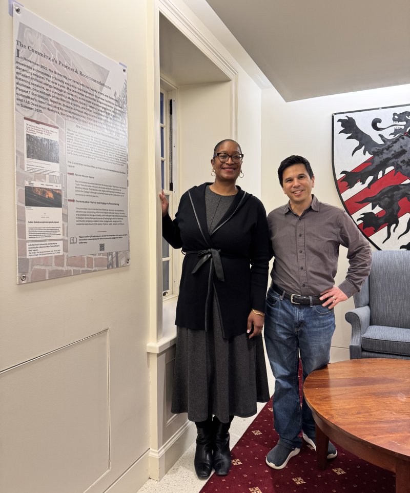 Two people standing and smiling indoors next to a wall display and a large crest.