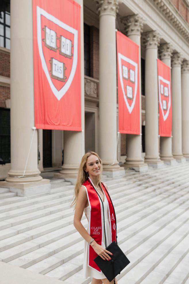 Lexi standing on the steps of Widener Library with her cap in her hands and dressed in graduation regalia.