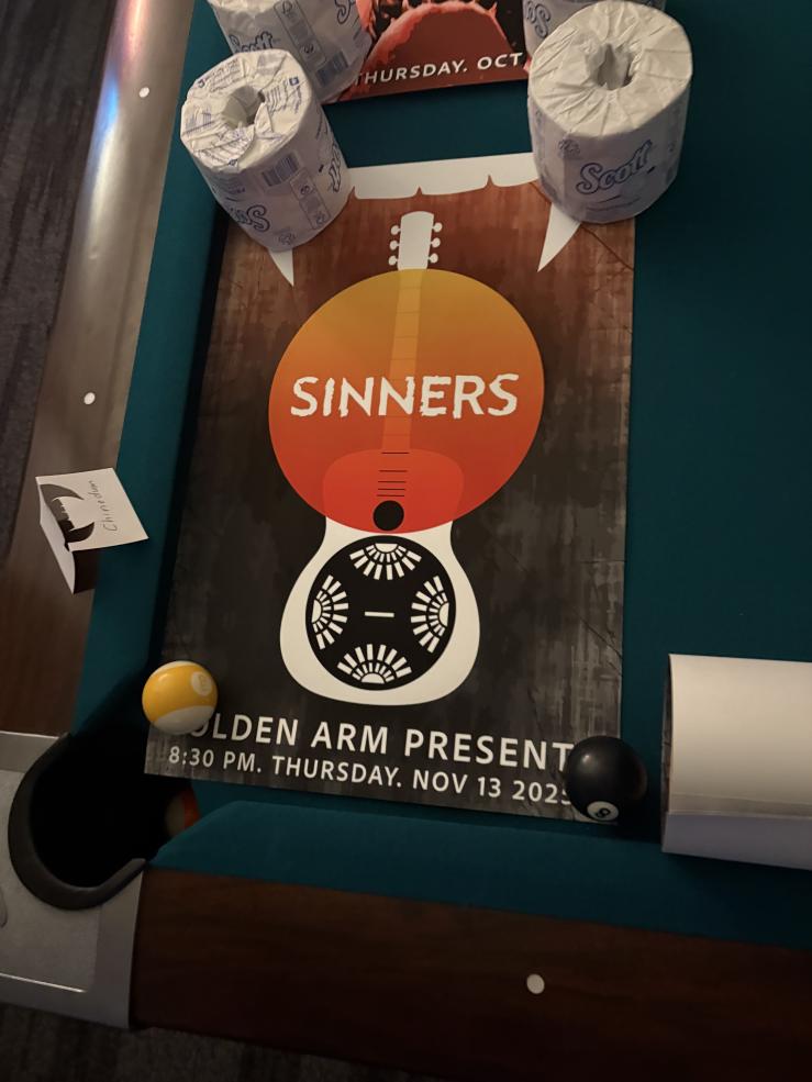 A poster held down by toilet paper roles with a guitar, an orange circle with the movie titled "Sinners" overlayed, and words reiterating the name of the theater and the date and time of the screening.