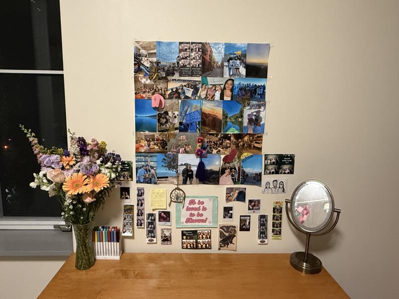 A student dorm desk with a vase of flowers on the left side, a mirror on the right side, and dozens of pictures on the wall.