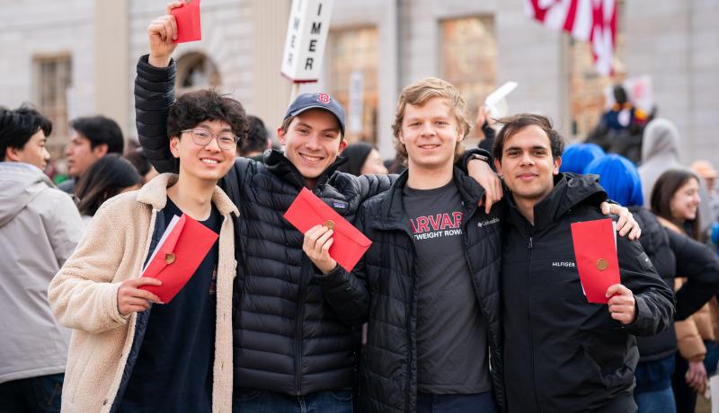 Four smiling students with arms around each other hold up red envelopes in a crowded campus courtyard.