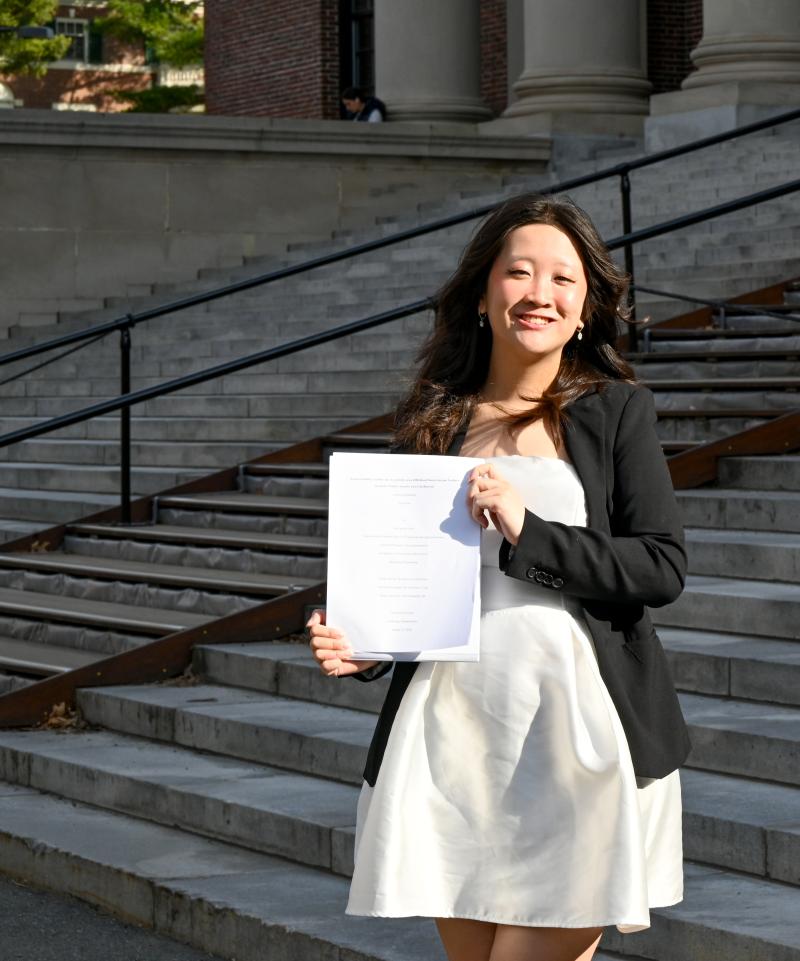 Lani Tran '26 standing on the steps of Widener Library holding up her thesis papers.