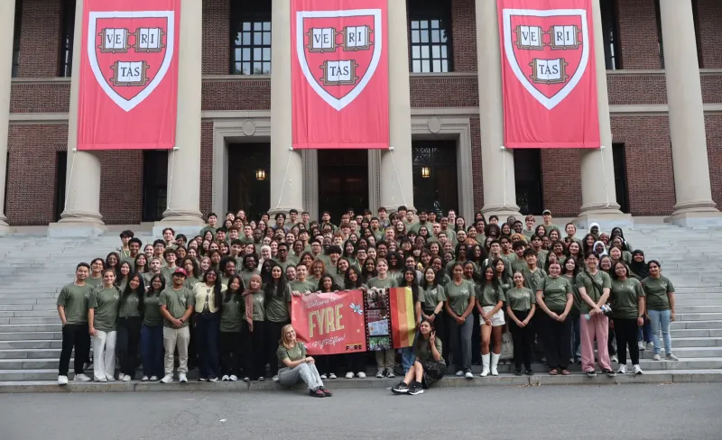 A wide group photograph of over a hundred college students standing on the grand stone steps of Widener Library, framed by massive white columns. Hanging between the columns are three large crimson banners featuring the Harvard University "VE RI TAS" crest. Every student wears a matching olive-green t-shirt. In the front center, two students sit on the ground while others hold a bright pink banner with playful drawings that reads "Welcome to FYRE FYREflies!"
