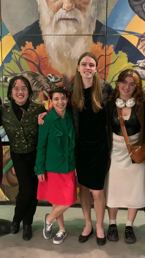 For people stand in front of a mural dressed in formal attire. The girl in the center wears a green jacket over a pink dress.