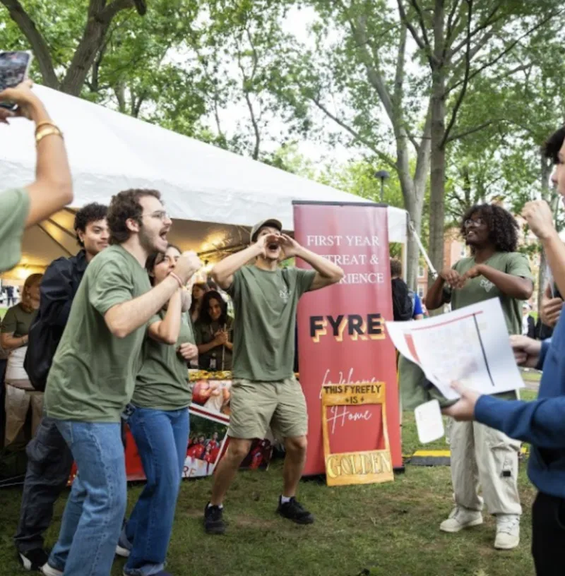 A vibrant outdoor photo of a group of diverse college student leaders cheering enthusiastically on a grassy campus lawn. They are wearing matching olive-green t-shirts. In the center, a student cups his hands around his mouth to shout a welcome chant. Behind them, a large white tent and a maroon vertical banner reading "FIRST YEAR RETREAT & EXPERIENCE FYRE" are visible.