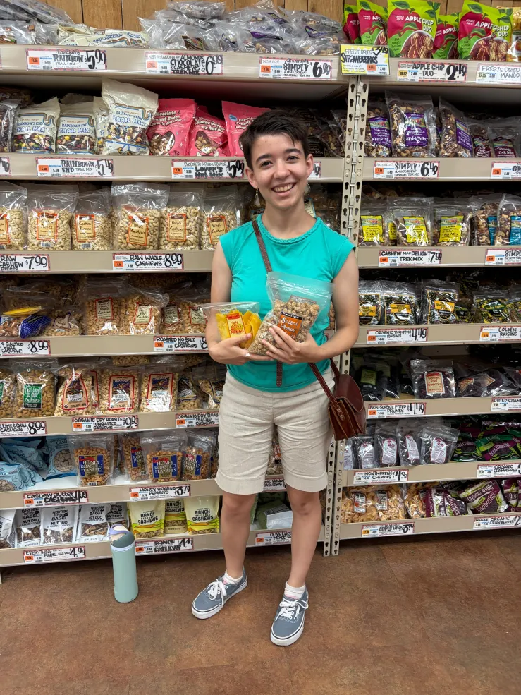 A student stands in front of a shelf of dried fruit and nuts at a grocery store. She is holding a bag of cashews and a bag of dried mangoes.