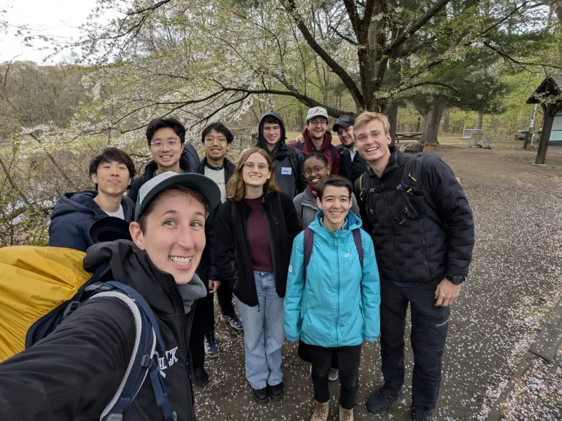 A group of students and community members stands in front of a budding spring tree in hiking gear