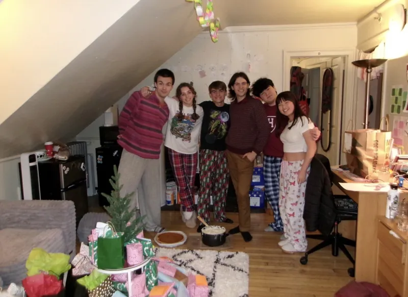 Six people standing together in a dorm common room, with presents and a Christmas tree in front