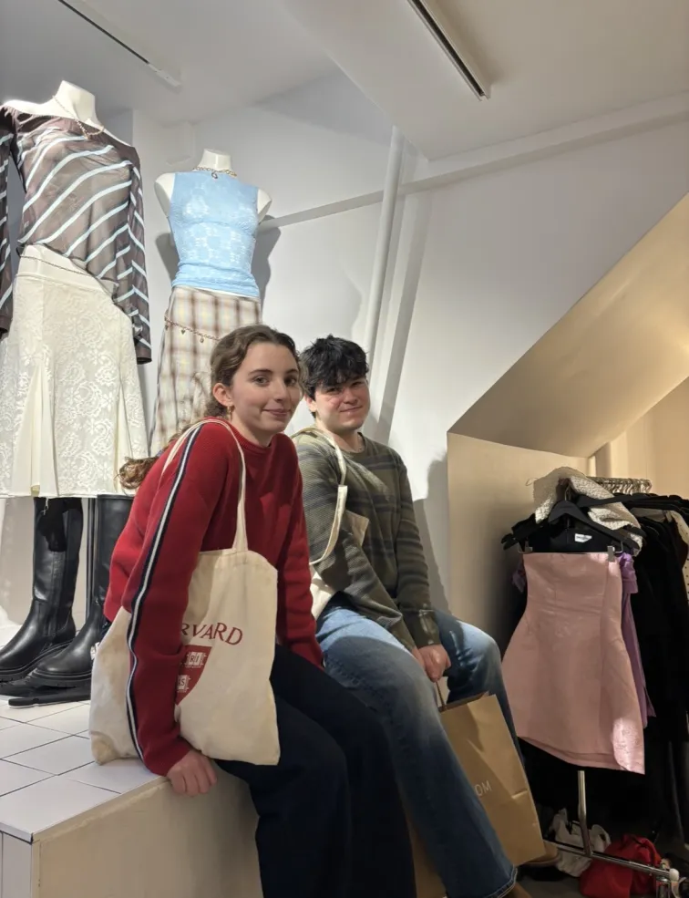 Two people sitting in a clothing store, with a rack of dresses next to them