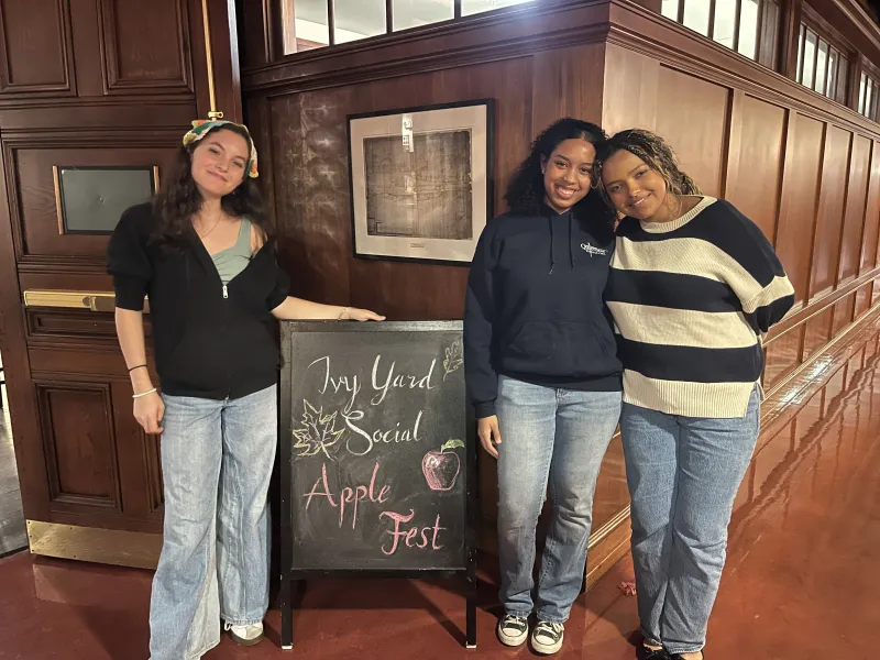 Three women standing next to a chalkboard reading "Ivy Yard Social Apple Fest"