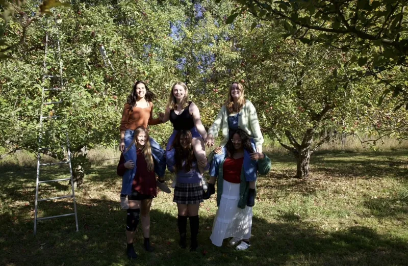 Three friends giving piggyback rides to three other friends in an apple orchard.