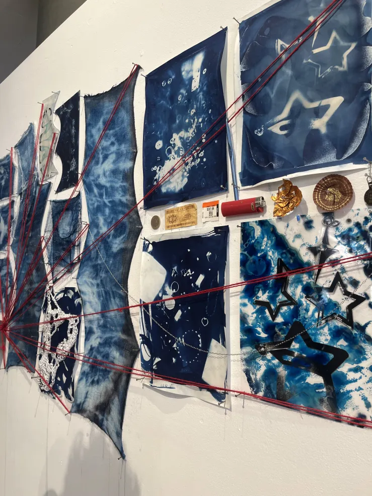Art display placed on wall. Blue and white fabrics connected by red string. Smaller trinkets: store tags, a lighter, and wrappers are placed in between fabrics.