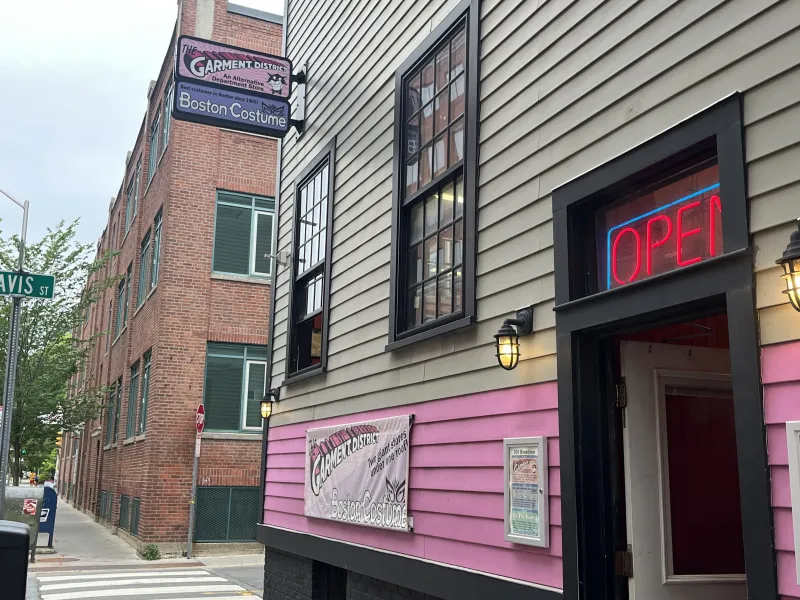 Gray and pink building with lamp lights and a neon "OPEN" sign above the door. Two signs protrude from the building, saying "The Garment District" and "Boston Costume" 
