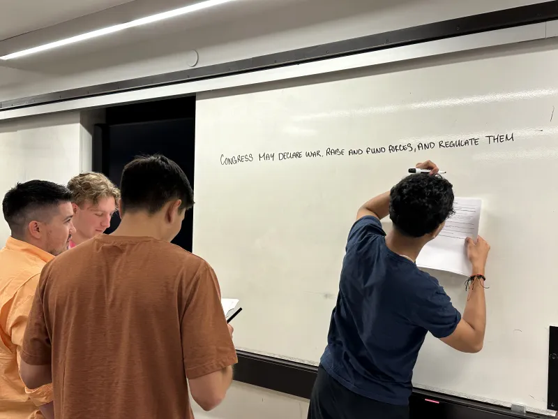 A group of four students surrounding a whiteboard, with one student writing on it with a marker.