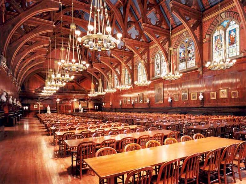 The interior of Annenberg Hall. Many rows of long tables sit within a long hall decorated with chandeliers and stained-glass windows.